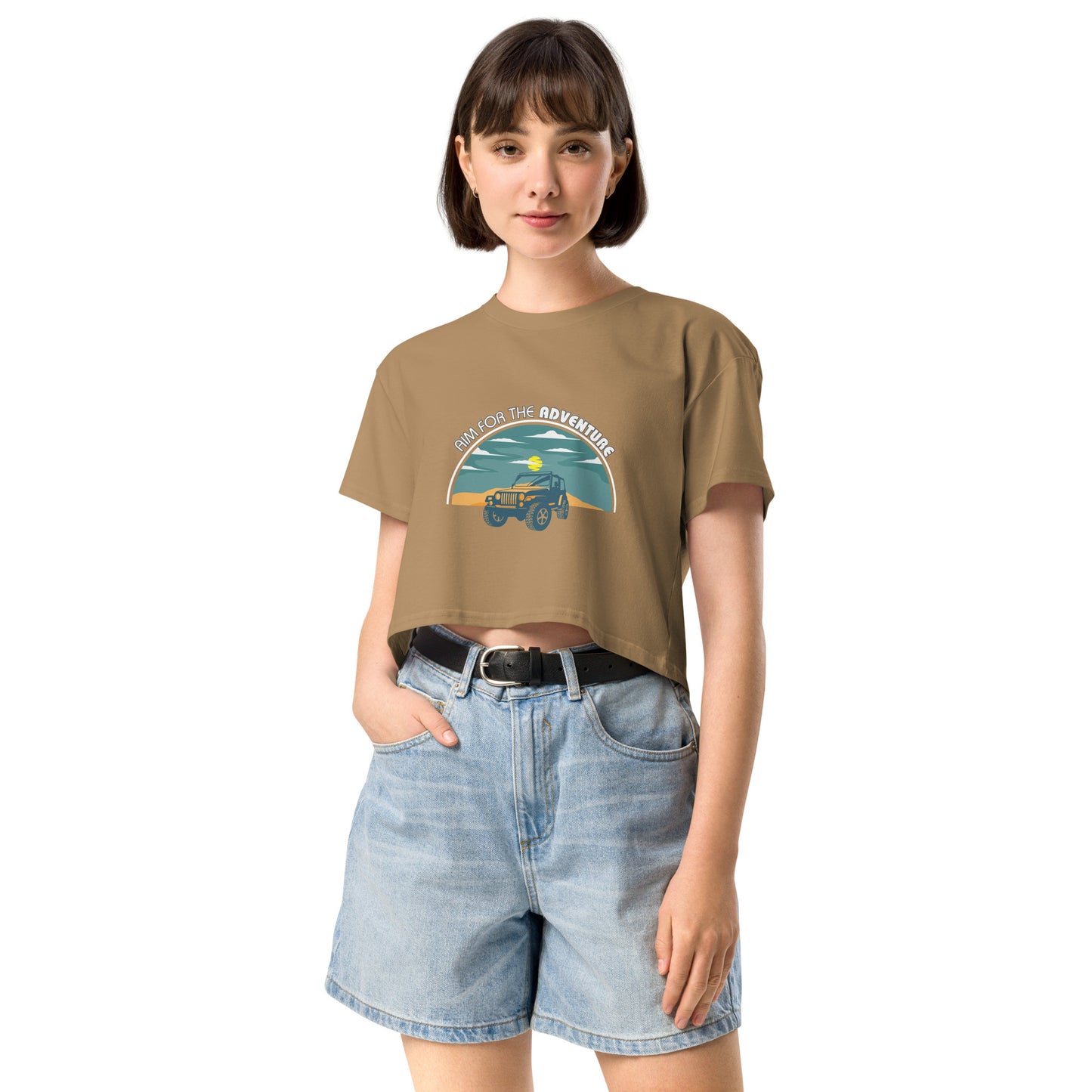 Content young woman wearing a camel crop top with a vintage car and 'AIM FOR THE ADVENTURE' graphic, paired with light denim shorts for a laid-back, adventurous vibe.