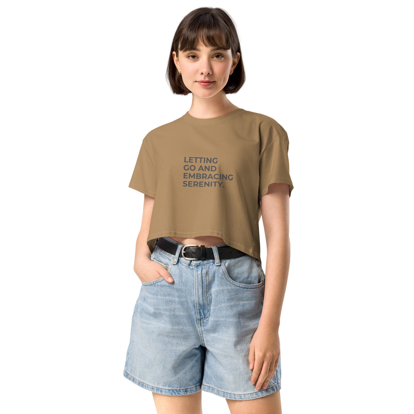 A young woman in a camel color cropped t-shirt with the text "Letting Go and Embracing Serenity" stands confidently with her hands in the pockets of her high-waisted denim shorts. She has a short bob haircut and is smiling slightly, creating a relaxed and serene vibe.