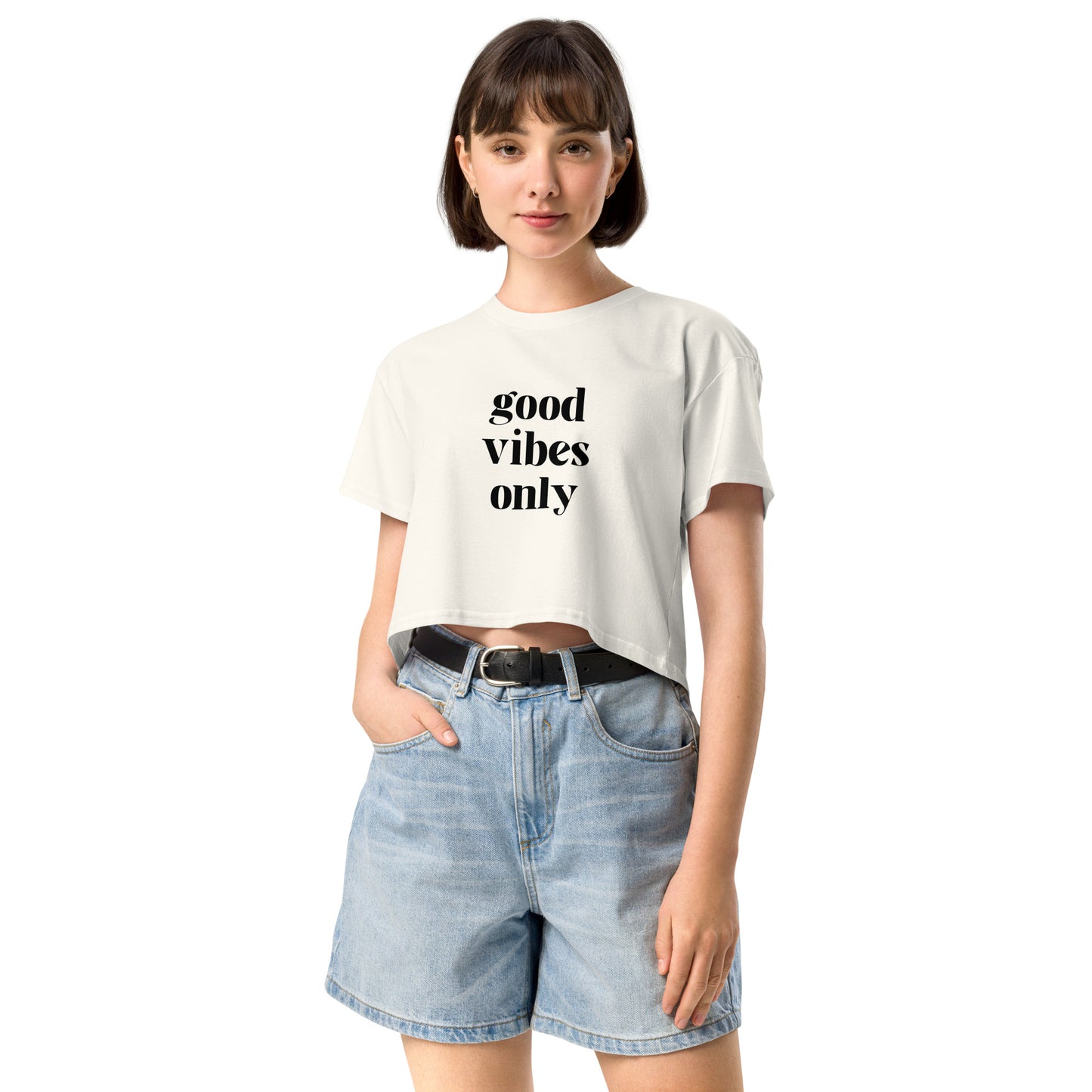 Content woman in a ecru crop top with 'good vibes only' text, coupled with light blue denim shorts, conveying a laid-back style.