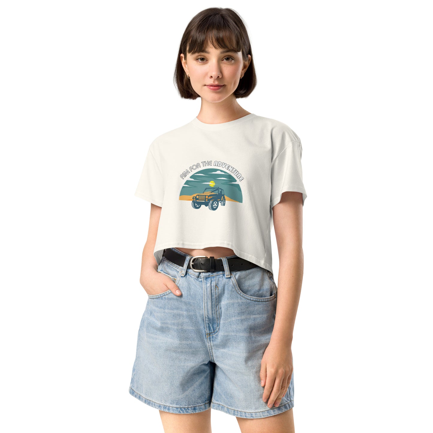 Content young woman wearing a ecru crop top with a vintage car and 'AIM FOR THE ADVENTURE' graphic, paired with light denim shorts for a laid-back, adventurous vibe.