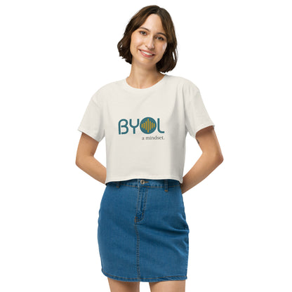 A young woman with short brown hair wearing a ecru white "BYOL: a mindset" crop top, smiling and standing with her hands on her hips. The crop top features the "BYOL" logo in teal and yellow on the front and is available in multiple colors (black, brown, white, light blue, plus more) and sizes (XS/SM - M/L). The "be you out loud" logo is displayed at the top.