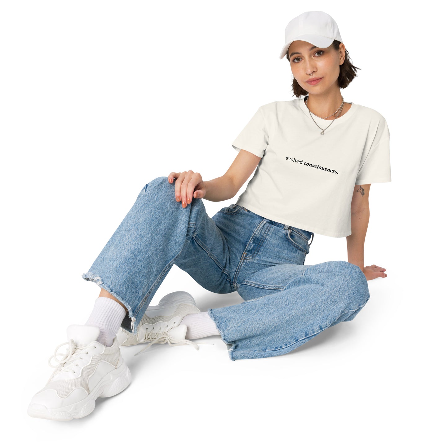 Woman wearing a ecru cropped t-shirt with the phrase "evolved consciousness" printed in black, sitting and posing against a clean background. Available in various colors: black, brown, pink, green, and white. Sizes range from XS to XL.