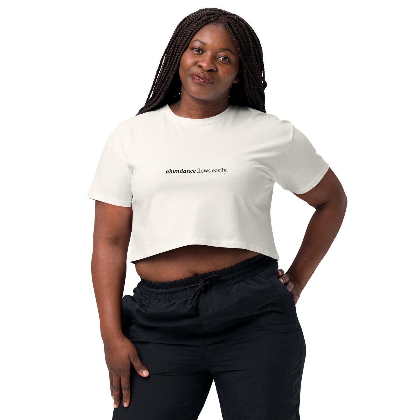 Woman wearing a ecru cropped t-shirt with the phrase "abundance flows easily" printed in black, smiling and posing against a clean background. Available in various colors: black, brown, pink, green, and white. Sizes range from XS to XL.