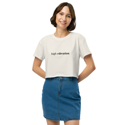 Woman wearing a orchid cropped t-shirt with the phrase "high vibration" printed in black, smiling and posing against a clean background. Available in various colors: black, brown, pink, green, and white. Sizes range from XS to XL.