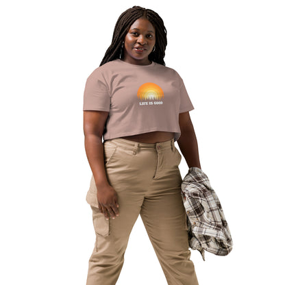 Woman in a hazy pink crop top with a "Life is Good" sunset graphic, pairing it with khaki pants and a casual jacket.