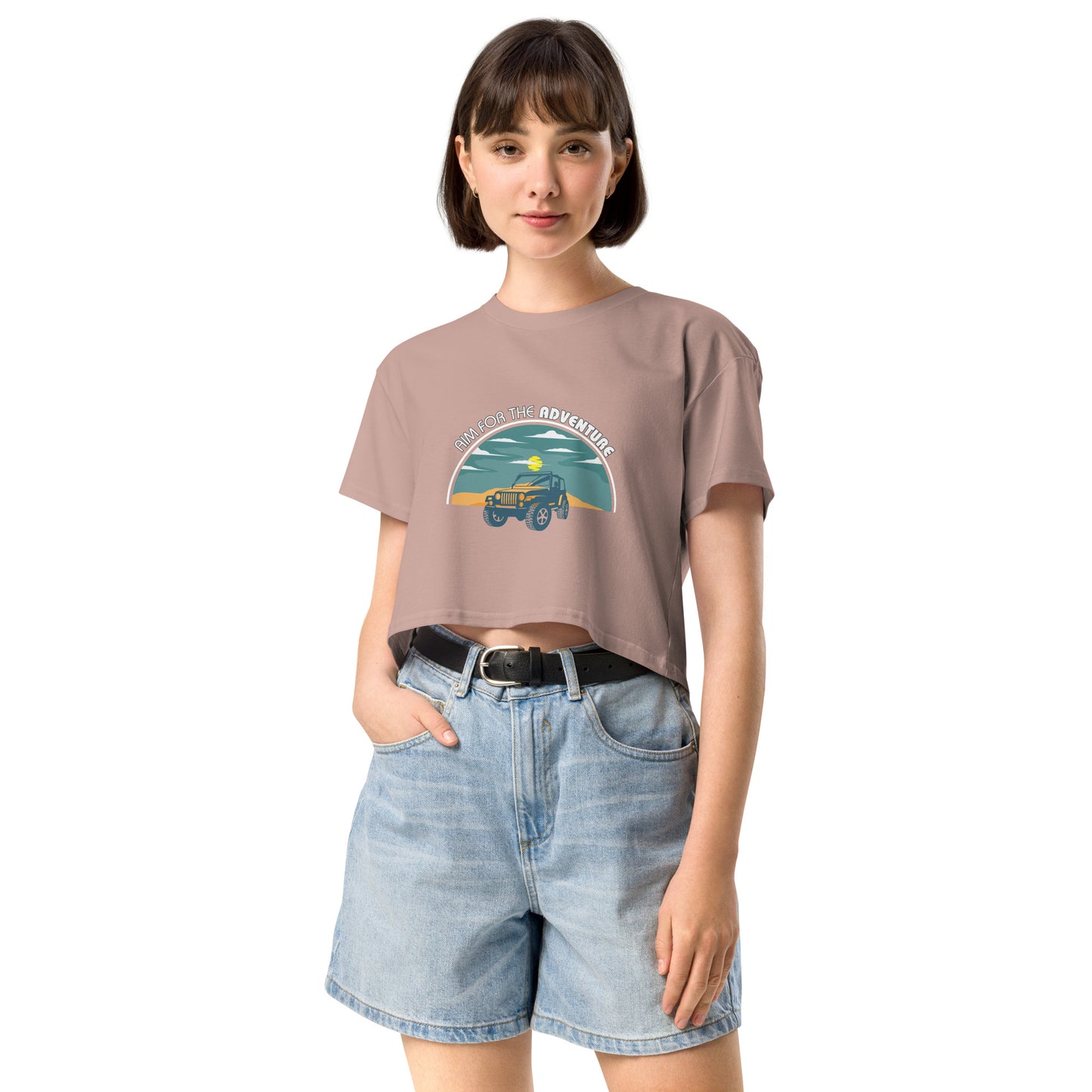 Content young woman wearing a hazy pink crop top with a vintage car and 'AIM FOR THE ADVENTURE' graphic, paired with light denim shorts for a laid-back, adventurous vibe.