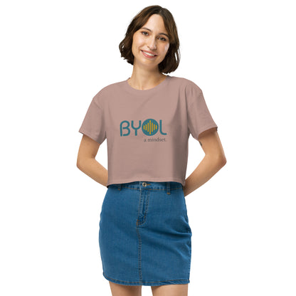 A young woman with short brown hair wearing a hazy pink "BYOL: a mindset" crop top, smiling and standing with her hands on her hips. The crop top features the "BYOL" logo in teal and yellow on the front and is available in multiple colors (black, brown, white, light blue, plus more) and sizes (XS/SM - M/L). The "be you out loud" logo is displayed at the top.