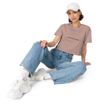 Woman wearing a hazy pink cropped t-shirt with the phrase "manifesting my reality" printed in black, sitting and posing against a clean background. Available in various colors: black, brown, pink, green, and white. Sizes range from XS to XL.