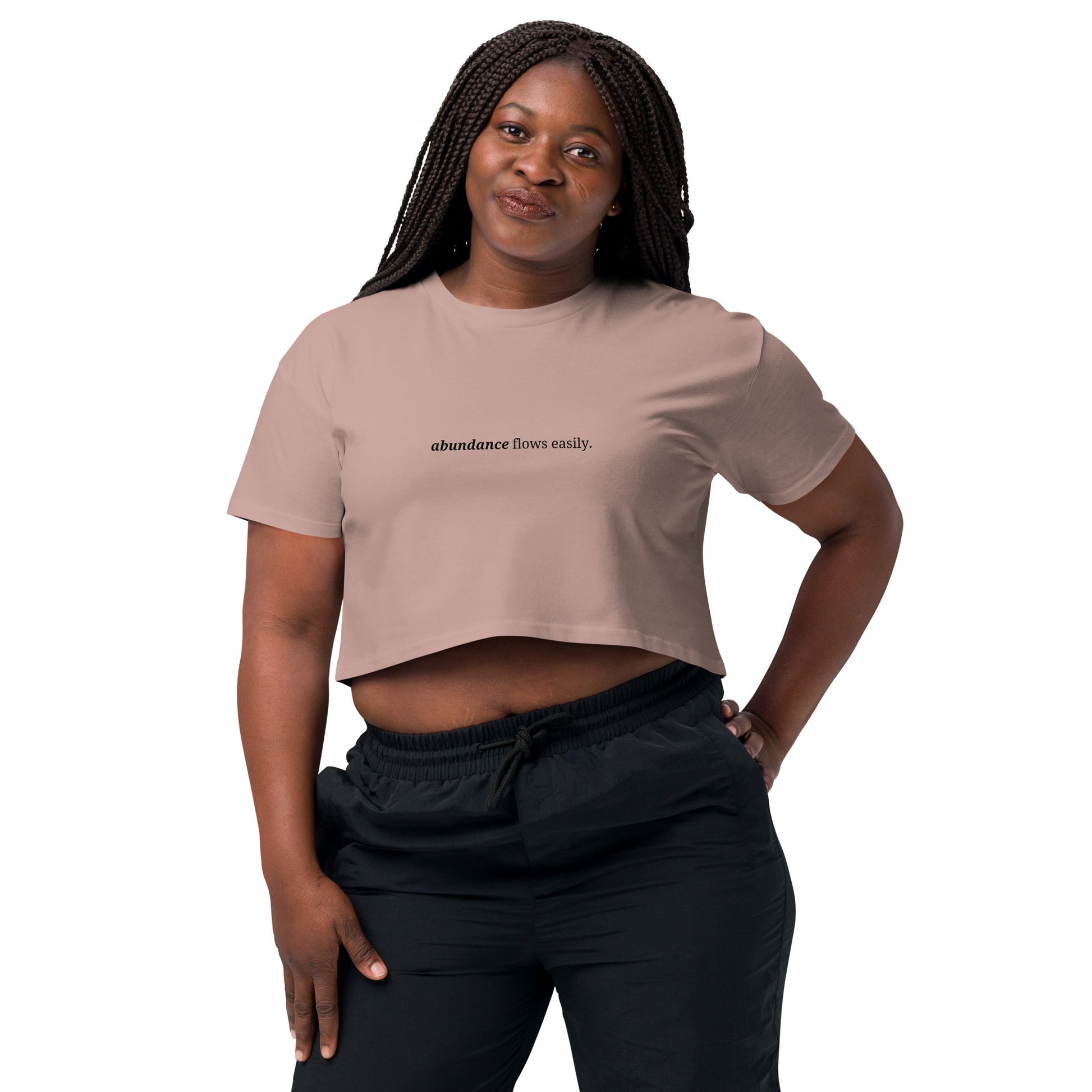 Woman wearing a hazy pink cropped t-shirt with the phrase "abundance flows easily" printed in black, smiling and posing against a clean background. Available in various colors: black, brown, pink, green, and white. Sizes range from XS to XL.