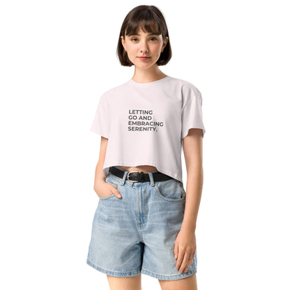 A young woman in a orchid cropped t-shirt with the text "Letting Go and Embracing Serenity" stands confidently with her hands in the pockets of her high-waisted denim shorts. She has a short bob haircut and is smiling slightly, creating a relaxed and serene vibe.