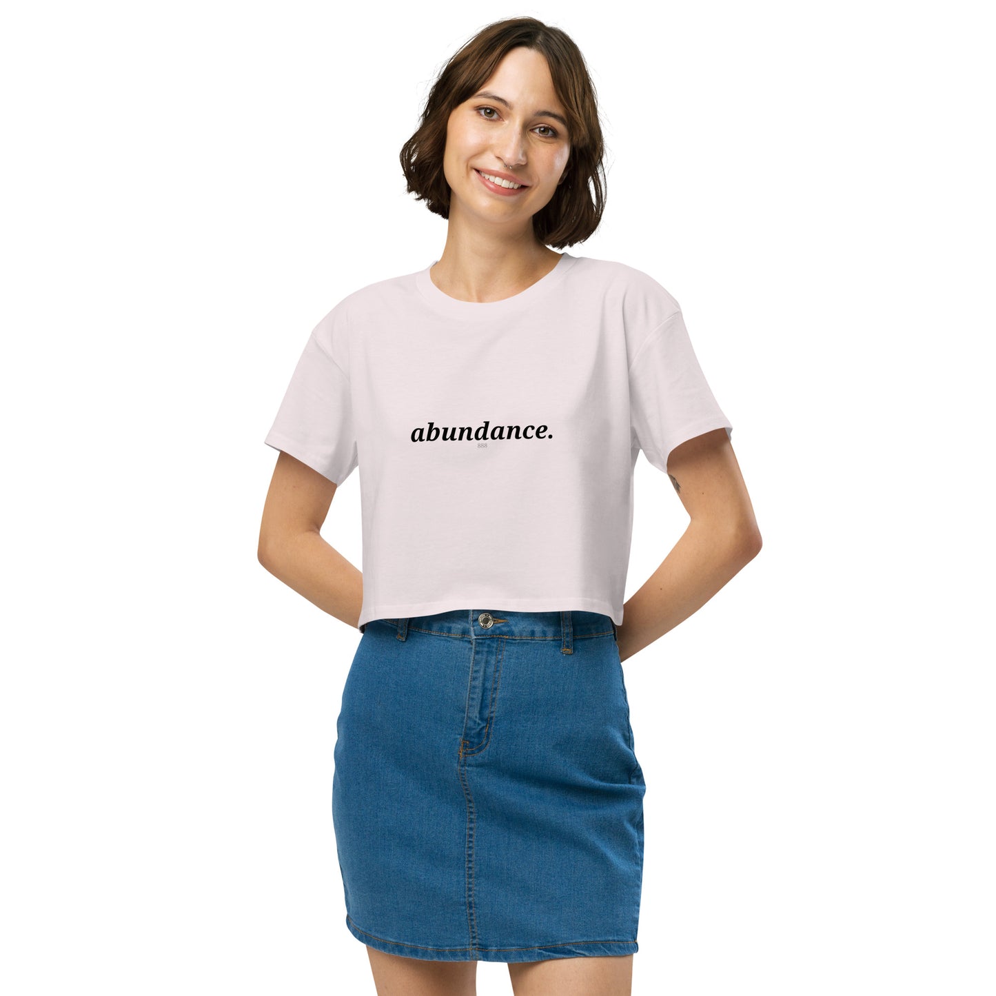 Woman wearing a orchid cropped t-shirt with the word "abundance" printed in black, smiling, and posing against a clean background. Available in various colors: black, brown, pink, green, and white. Sizes range from XS to XL.