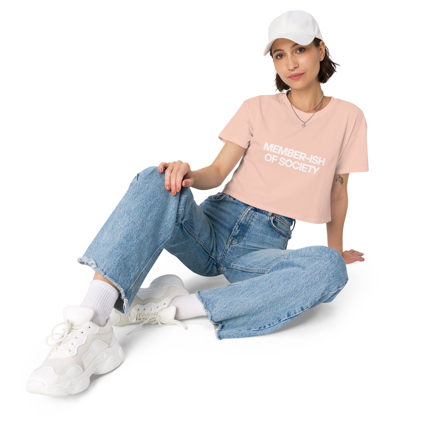 Casual and cool woman in a pale pink crop top with 'Member-ish of Society' text, paired with denim jeans, white sneakers, and a cap for a laid-back look.