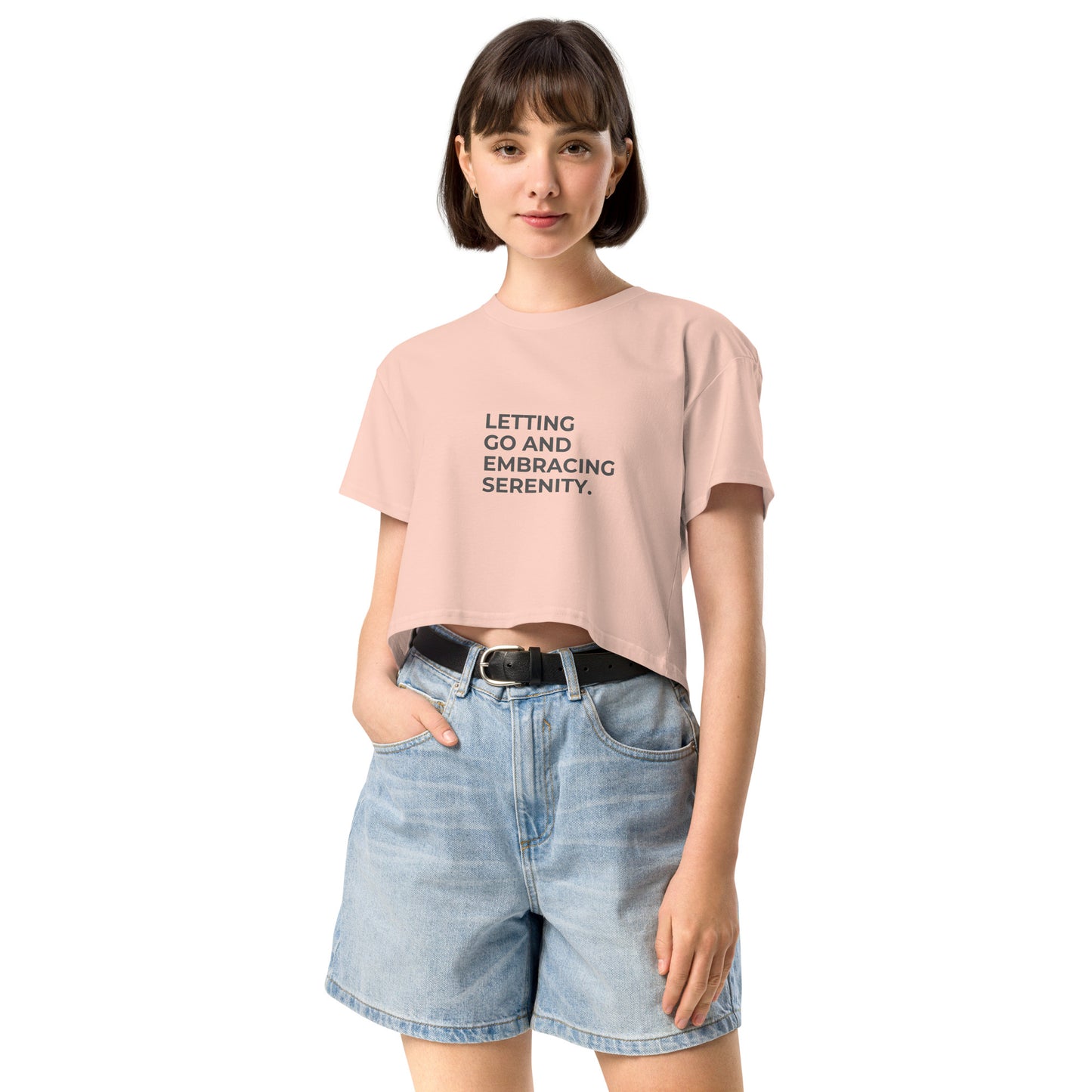 A young woman in a pale pink cropped t-shirt with the text "Letting Go and Embracing Serenity" stands confidently with her hands in the pockets of her high-waisted denim shorts. She has a short bob haircut and is smiling slightly, creating a relaxed and serene vibe.