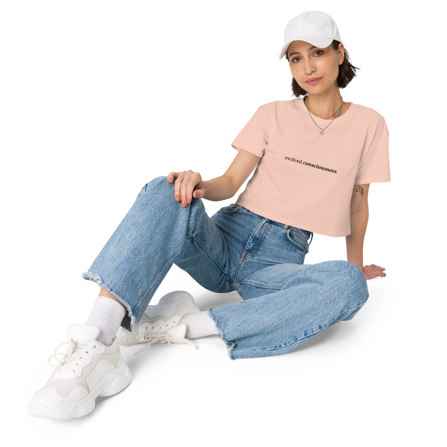 Woman wearing a pale pink cropped t-shirt with the phrase "evolved consciousness" printed in black, sitting and posing against a clean background. Available in various colors: black, brown, pink, green, and white. Sizes range from XS to XL.