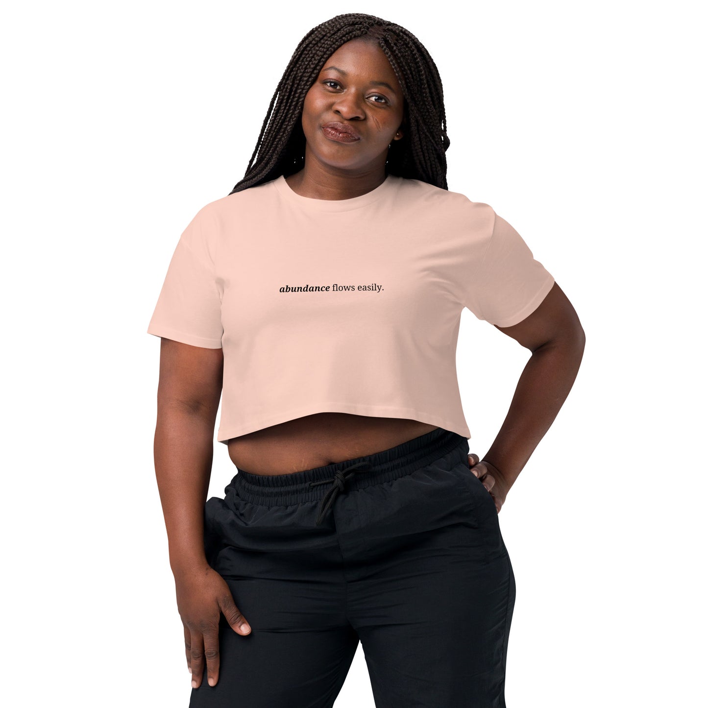 Woman wearing a pale pink cropped t-shirt with the phrase "abundance flows easily" printed in black, smiling and posing against a clean background. Available in various colors: black, brown, pink, green, and white. Sizes range from XS to XL.