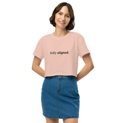 Woman wearing a pale pink cropped t-shirt with the phrase "fully aligned" printed in black, smiling and posing against a clean background. Available in various colors: black, brown, pink, green, and white. Sizes range from XS to XL.