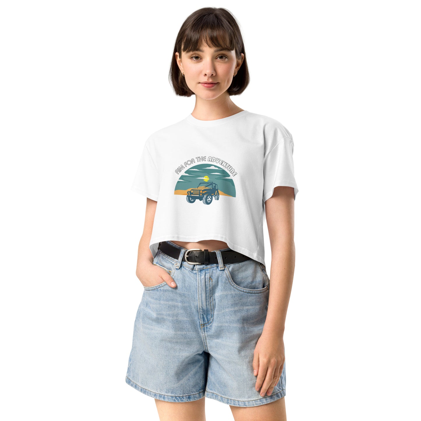 Content young woman wearing a white crop top with a vintage car and 'AIM FOR THE ADVENTURE' graphic, paired with light denim shorts for a laid-back, adventurous vibe.