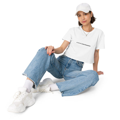 Woman wearing a white cropped t-shirt with the phrase "evolved consciousness" printed in black, sitting and posing against a clean background. Available in various colors: black, brown, pink, green, and white. Sizes range from XS to XL.