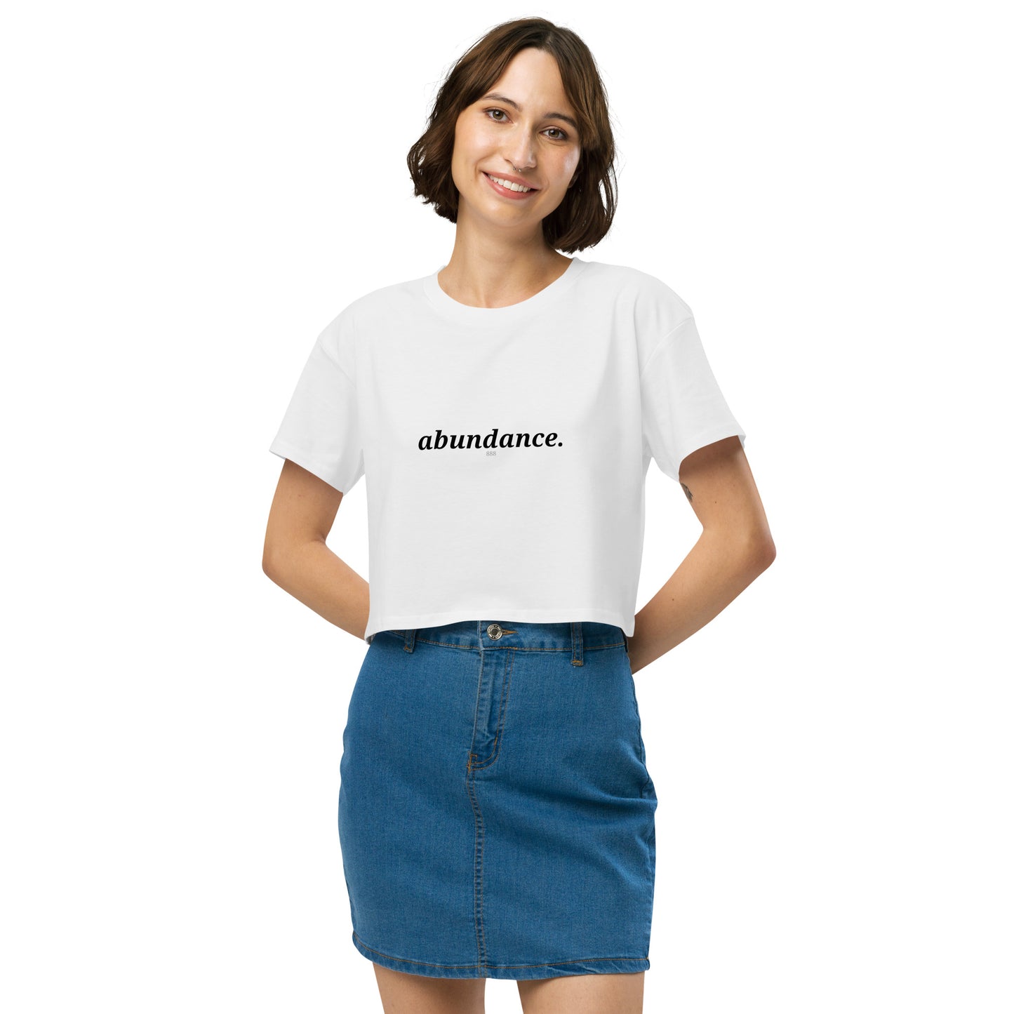 Woman wearing a white cropped t-shirt with the word "abundance" printed in black, smiling, and posing against a clean background. Available in various colors: black, brown, pink, green, and white. Sizes range from XS to XL.