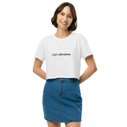 Woman wearing a white cropped t-shirt with the phrase "high vibration" printed in black, smiling and posing against a clean background. Available in various colors: black, brown, pink, green, and white. Sizes range from XS to XL.