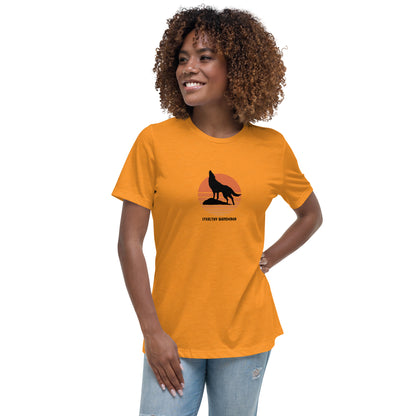 Woman wearing a heather orange bravery-themed relaxed t-shirt with a wolf graphic and 'Stealthy Wanderer' text in a sunset backdrop