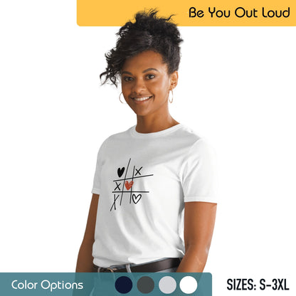 A woman with an updo hairstyle and hoop earrings is wearing a white T-shirt with a tic-tac-toe grid design that includes hearts and an "X" on the grid. She’s smiling and looking towards the camera with a backdrop featuring the campaign slogan "Be You Out Loud," a color palette, and available sizes from S to 3XL.